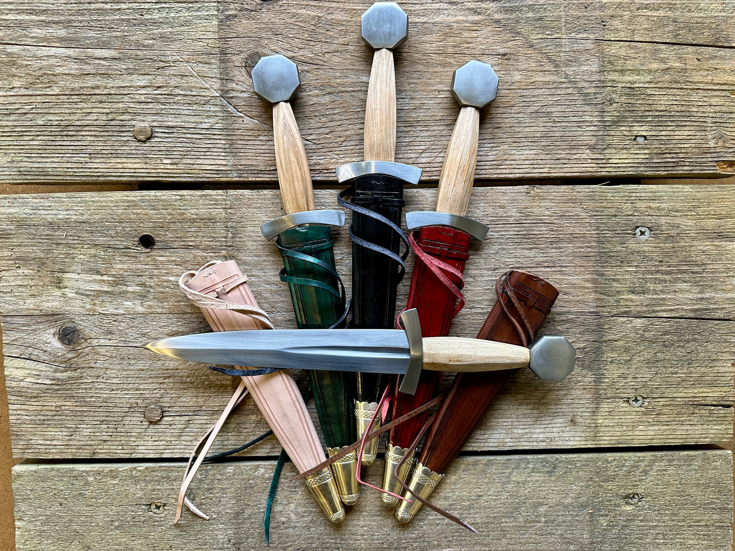 Late 13thC Quillon Dagger. Array of all scabbards in red, green, brown, black and natural with a knife on top.