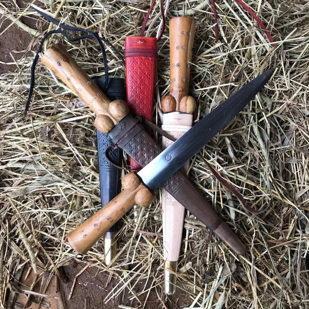 Three dot and circle bollock daggers with black, red, brown and natural scabbards on a bed of straw.