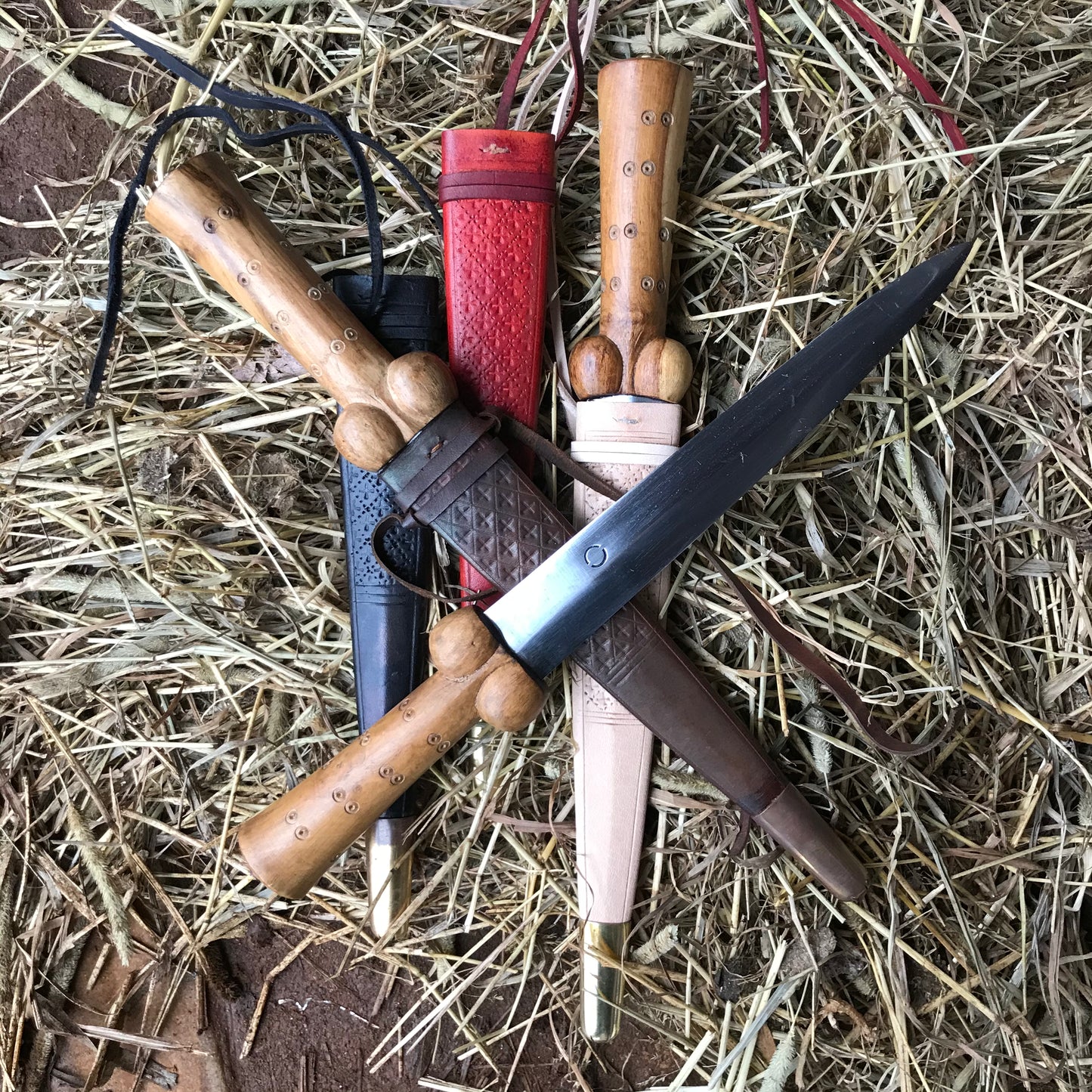 Three dot and circle bollock daggers with black, red, brown and natural scabbards on a bed of straw.