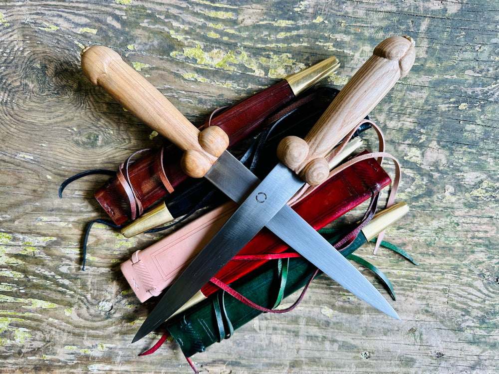 Two rude bollock daggers on a bed of scabbards in red, black, natural, brown and green