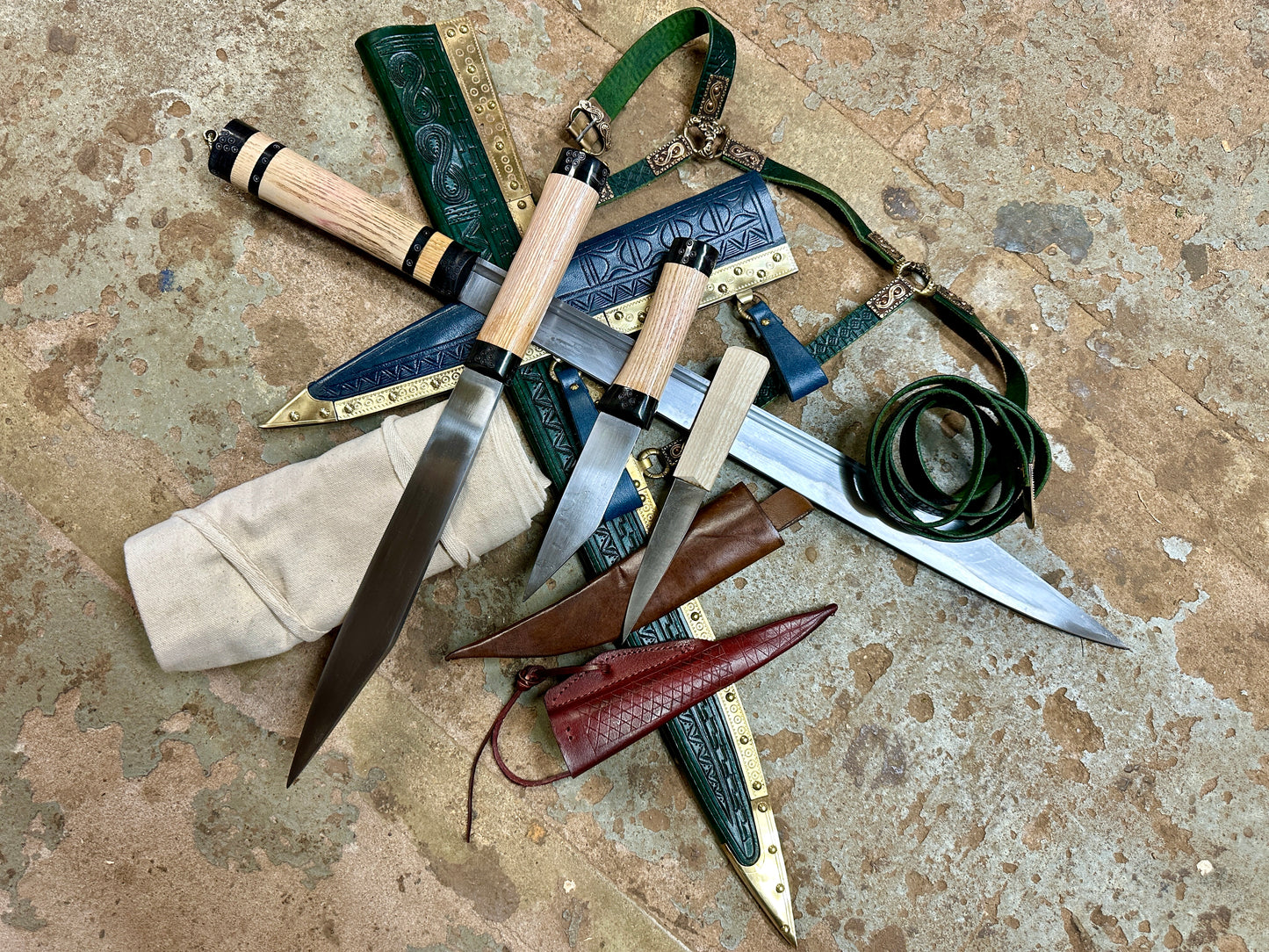 Bundle of Sword Seax, Langseax, Viking everyday carry and viking eating knife in a pile on their scabbards with a cotton sword bag rolled up next to them. 